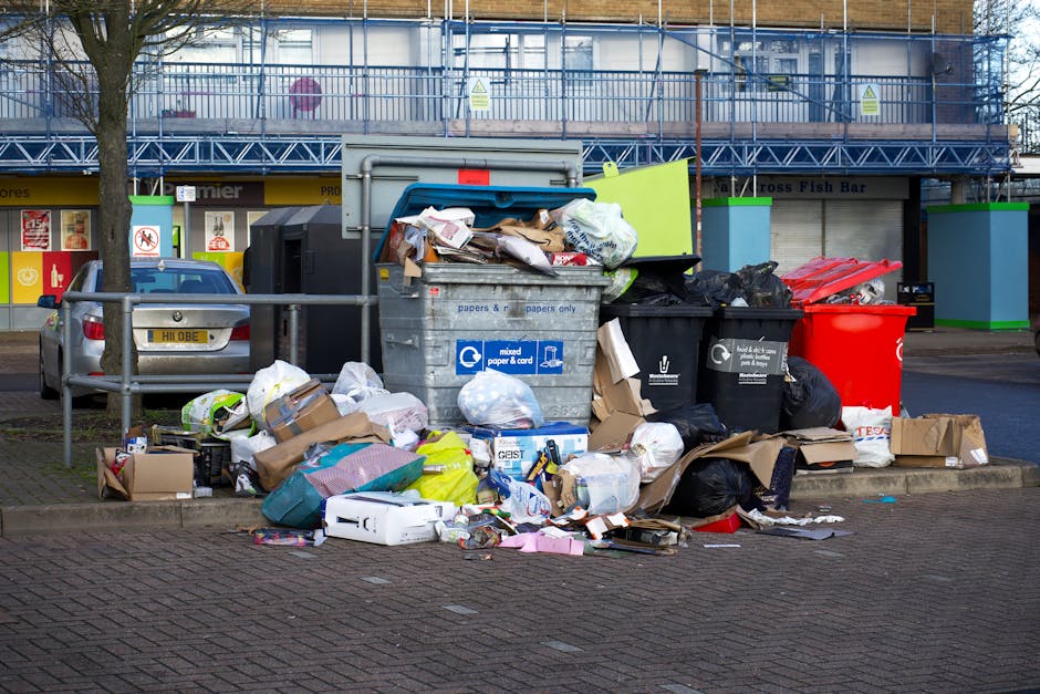 A black metal public rubbish bin situated on a cobblestone pavement in an urban setting, overflowing with various trash including paper and plastic packaging. The top of the bin is cluttered with used