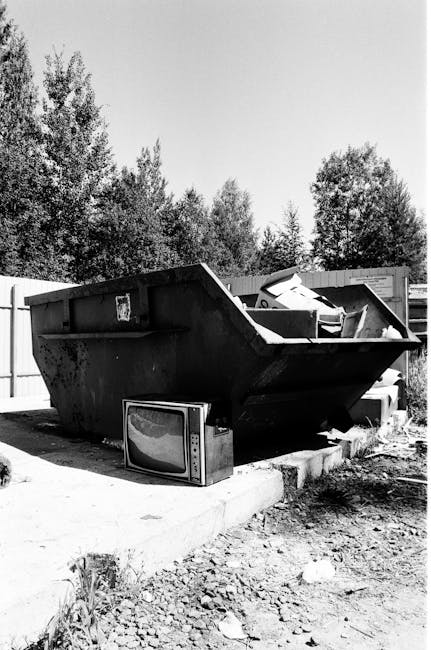 A large, black, metal skip bin is positioned on a concrete surface in an outdoor area, surrounded by a wooden fence and with trees and foliage visible in the background. The skip has a rectangular sha