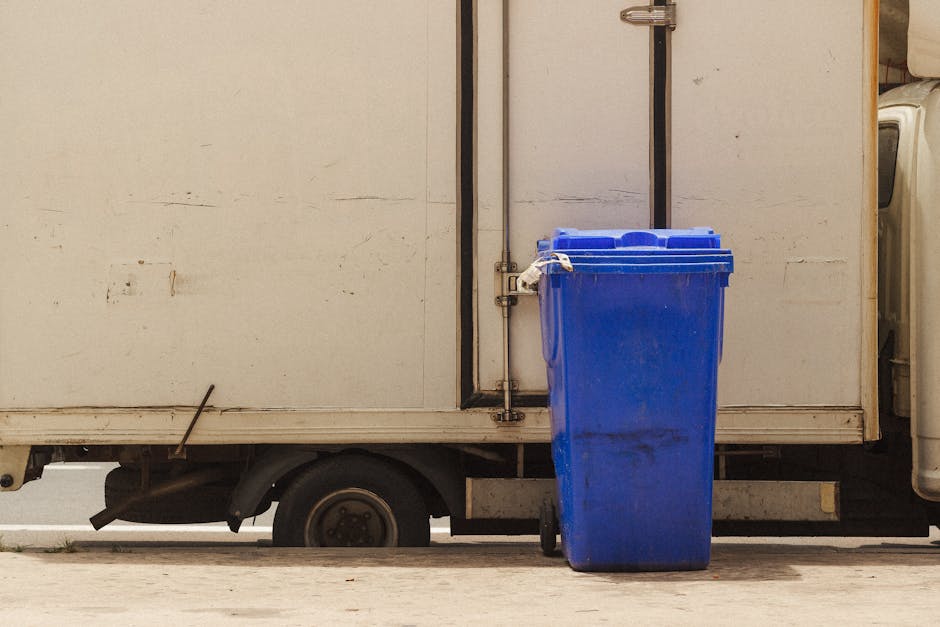 A large, black, metal skip bin is positioned on a concrete surface in an outdoor area, surrounded by a wooden fence and with trees and foliage visible in the background. The skip has a rectangular sha