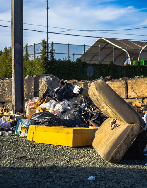 A pile of mixed waste materials is situated against a brick and concrete block wall, consisting of flattened cardboard boxes, plastic packaging, and a large woven fabric sack. Several cardboard boxes